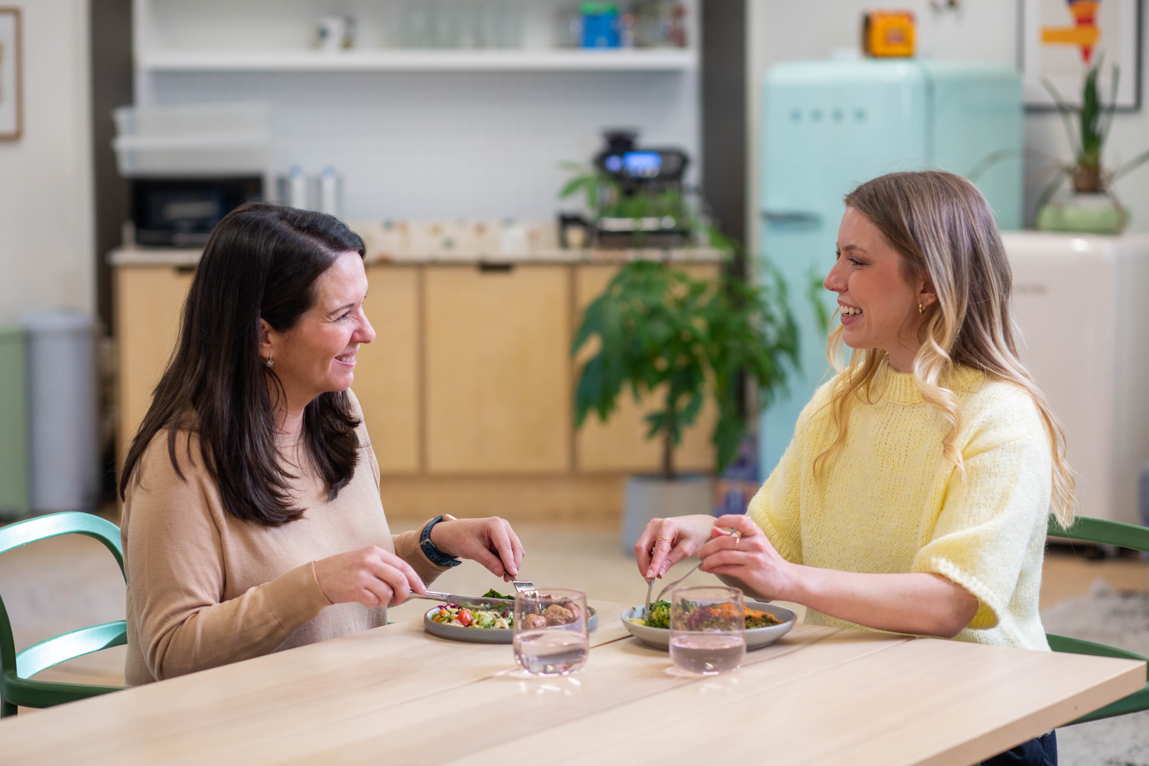 A woman sitting on a sofa at home, smiling while holding a plate of food, with houseplants in the foreground
